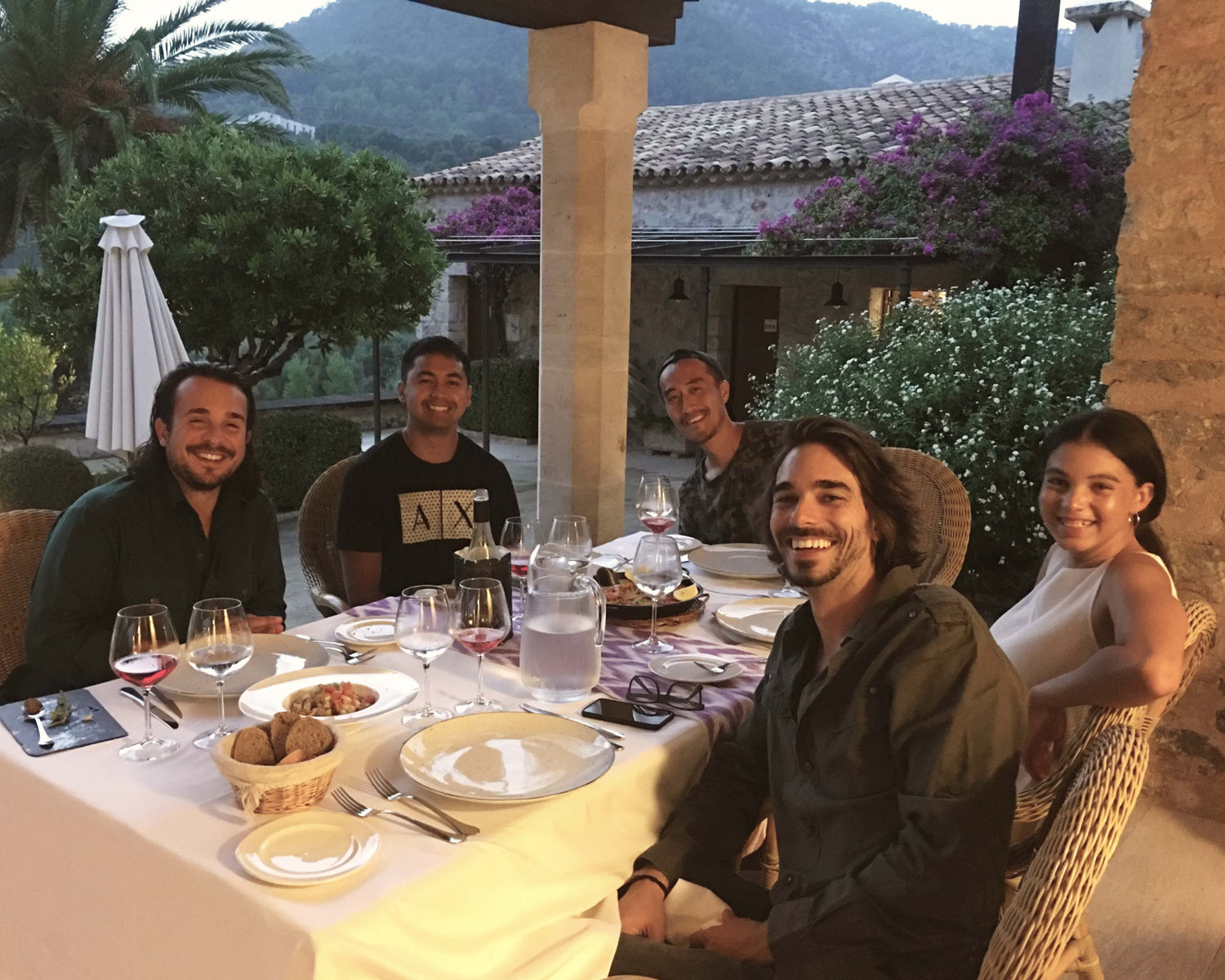 A group of five people sitting around a dinner table outdoors, smiling at the camera. The table is set with plates, glasses of wine, and food, with a relaxed atmosphere under a patio. The background features lush greenery, flowers, and a scenic view of mountains at dusk.