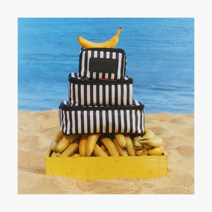 Set of Four / Poplin Stripe Black | Stack of Poplin Stripe Black compressible packing cubes arranged on a crate of bananas on a sandy beach, with a single banana placed on top